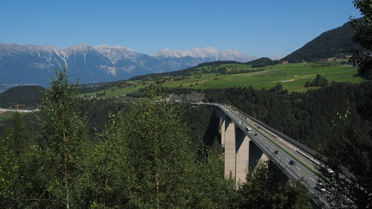A motorway viaduct on tall concrete piers crossing a forested alpine valley, snow-streaked peaks behind