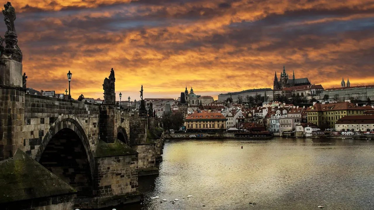 Charles Bridge over the Vltava at sunset, with Prague Castle and red-tiled roofs on the far bank
