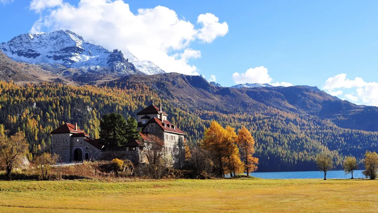 A stone manor beside an alpine lake in autumn, golden larch forest and a snow-capped peak above