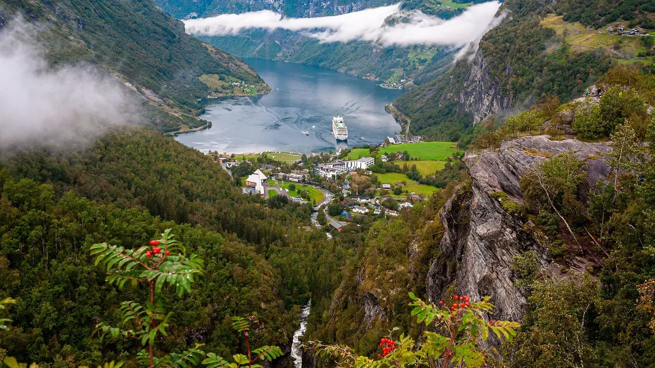 A Norwegian fjord seen from a clifftop, a cruise ship at anchor and a village on the green valley floor