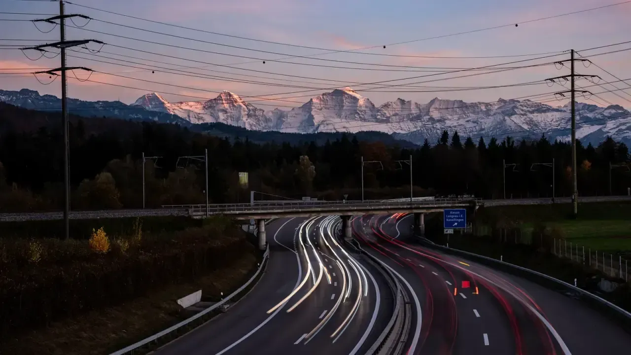 A Swiss motorway at dusk with traffic light trails and the snow-capped Alps behind